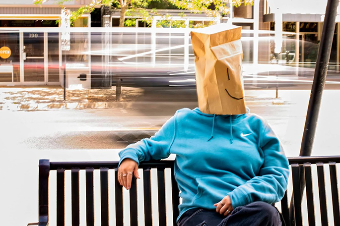 Person in blue hoodie, sitting on a bench, wearing a paper bag with a smiley face on their head