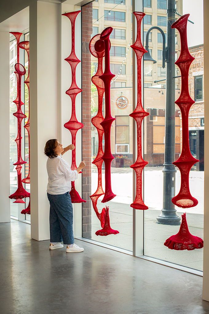 Woman in a white shirt and blue pants looking up at hanging red crochet