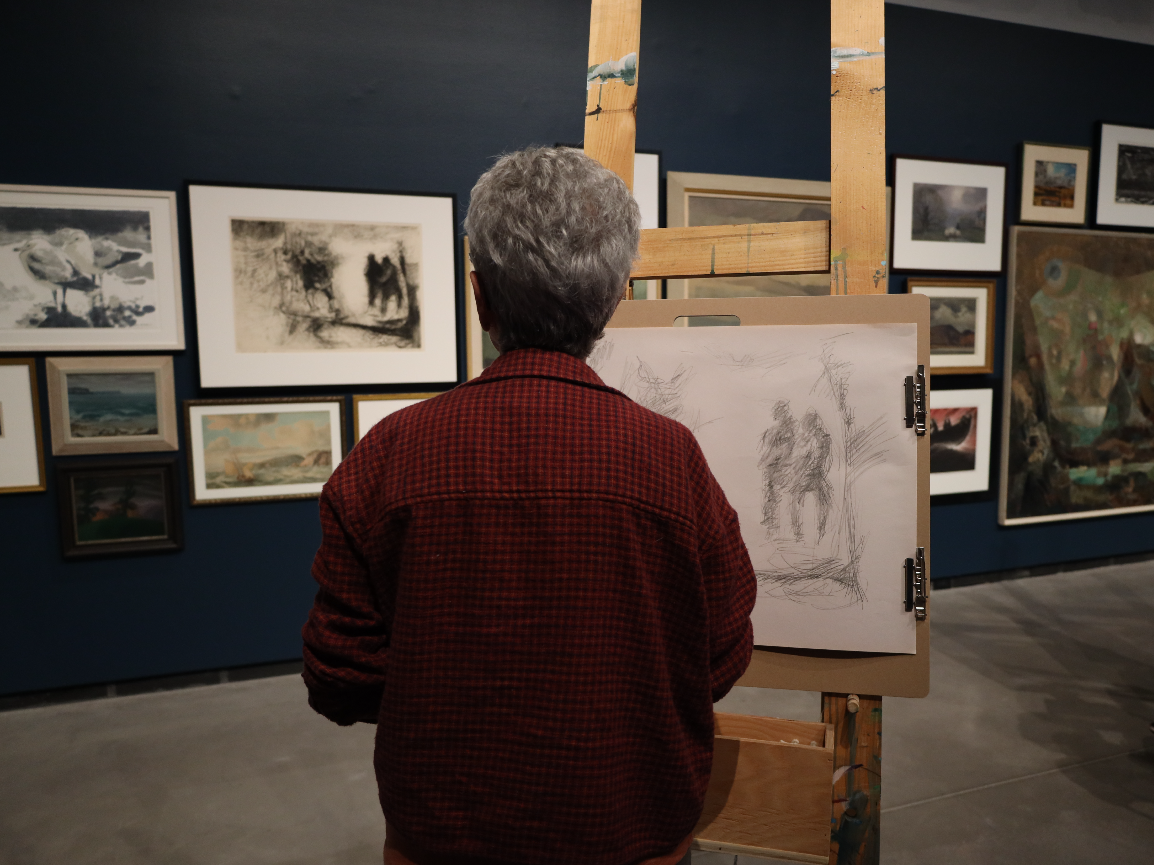 Visitor sketching at an easel in an art gallery surrounded by framed artwork.