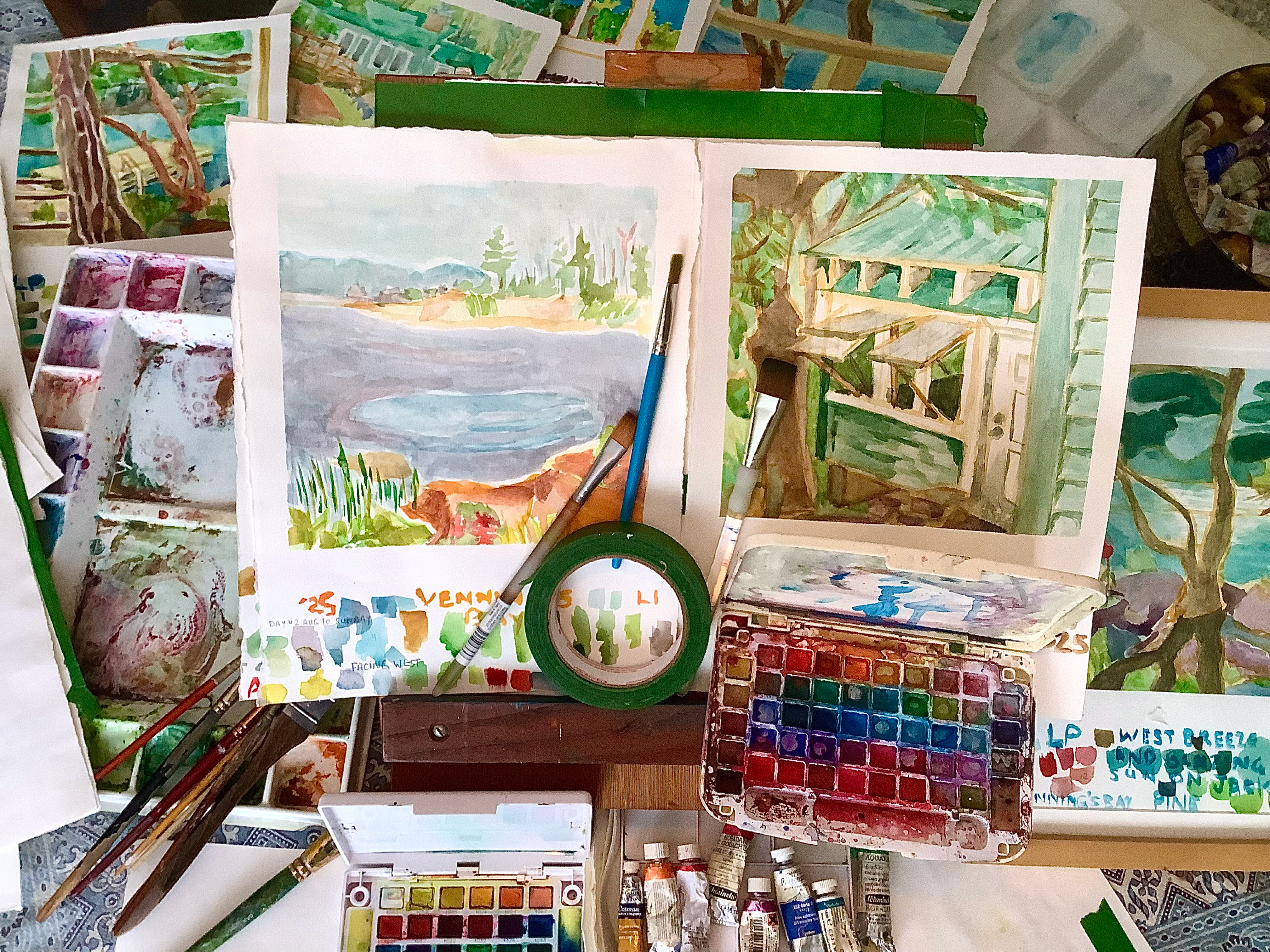 Watercolor paintings, palettes, brushes, and paint tubes arranged on a tabletop around two landscape artworks.