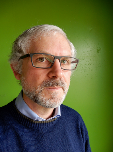 Man with short grey hair and glasses standing in front of a bright green backdrop. 