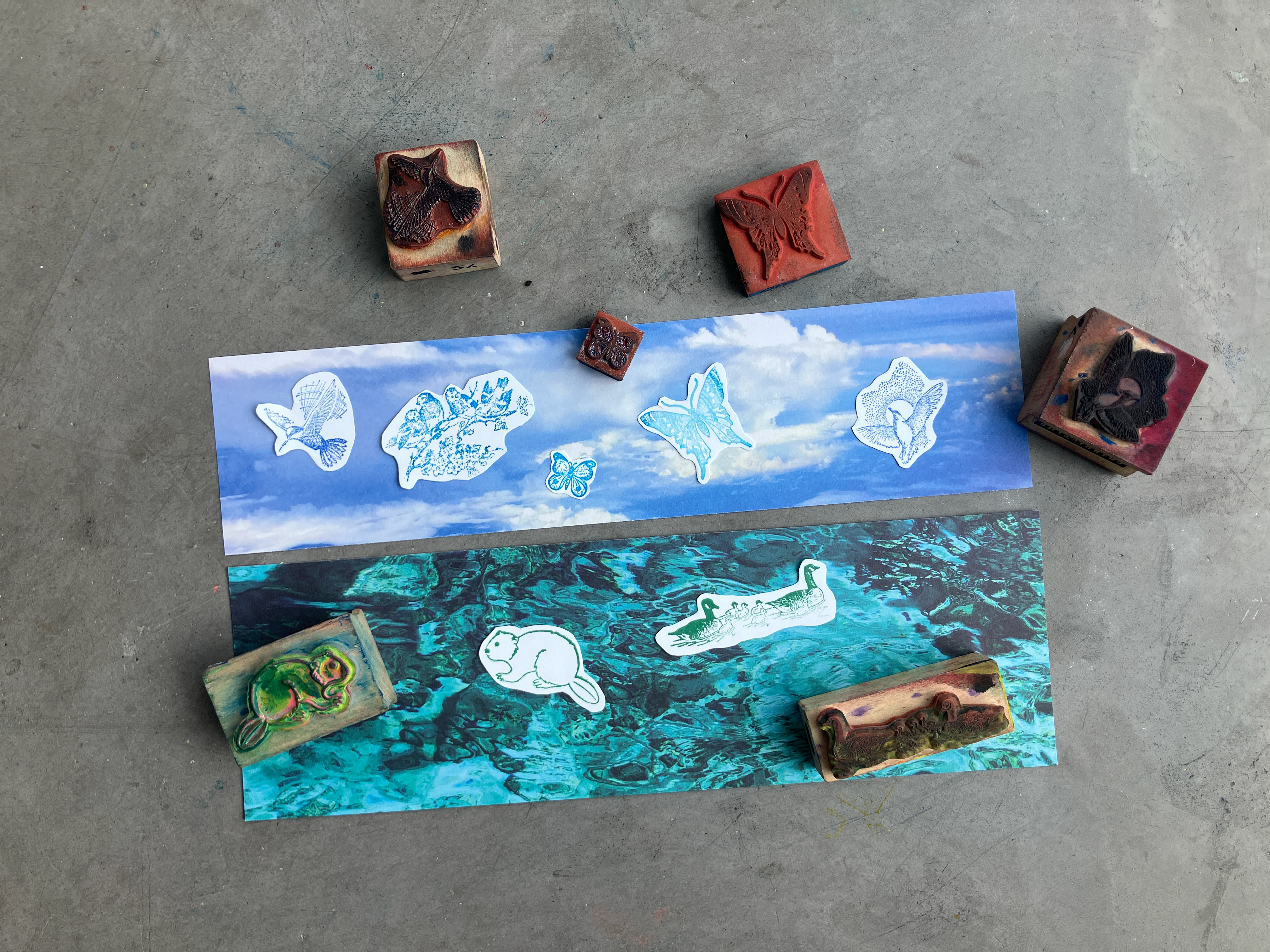 Two photo strips with sky and water backgrounds, decorated with butterfly and animal cut-outs, surrounded by inked stamps.