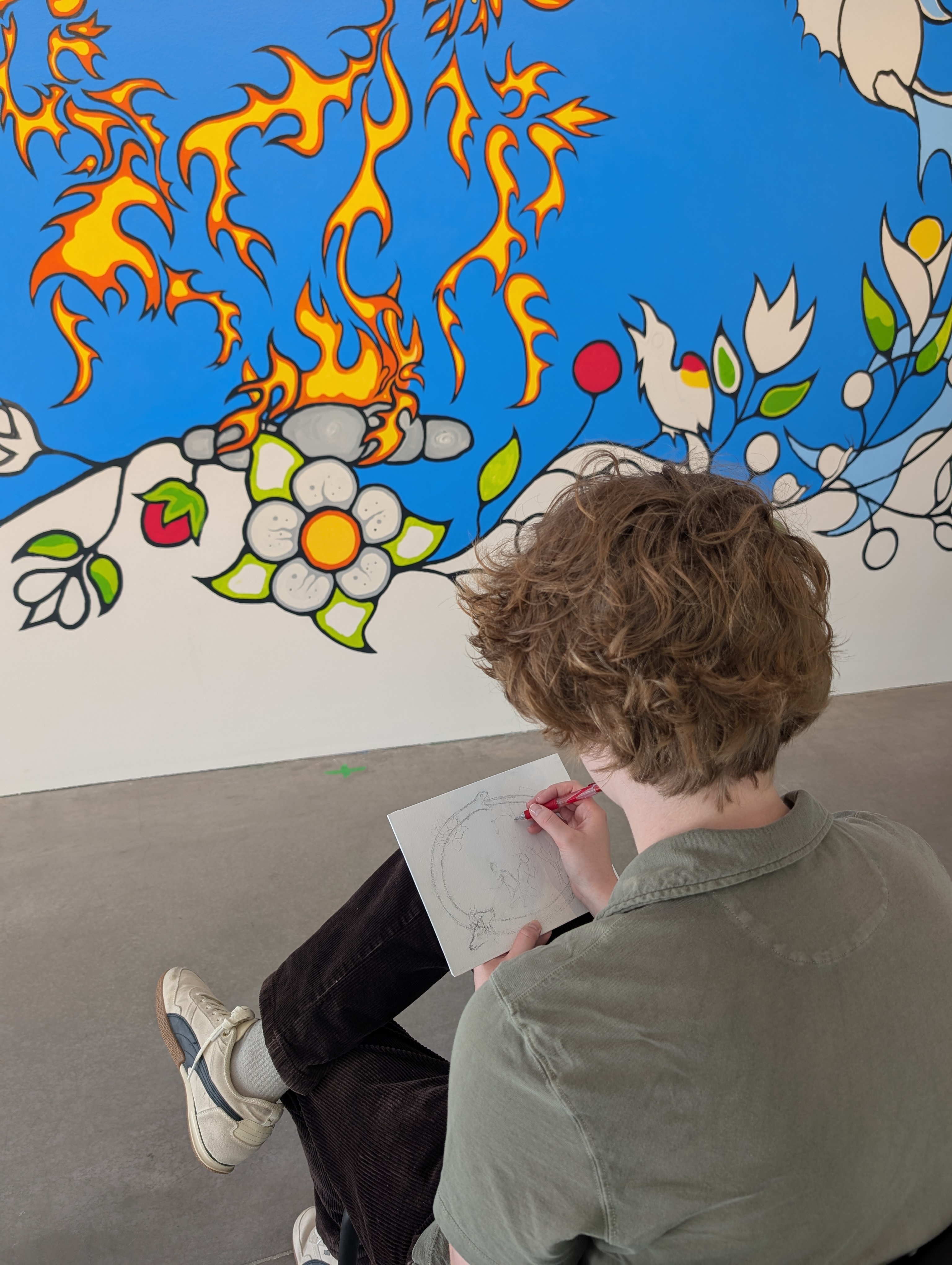 A person with short, curly brown hair, and wearing an army green shirt sketches in front of John Williams' mural in the lecture theatre of the Judith & Norman Alix Art Gallery