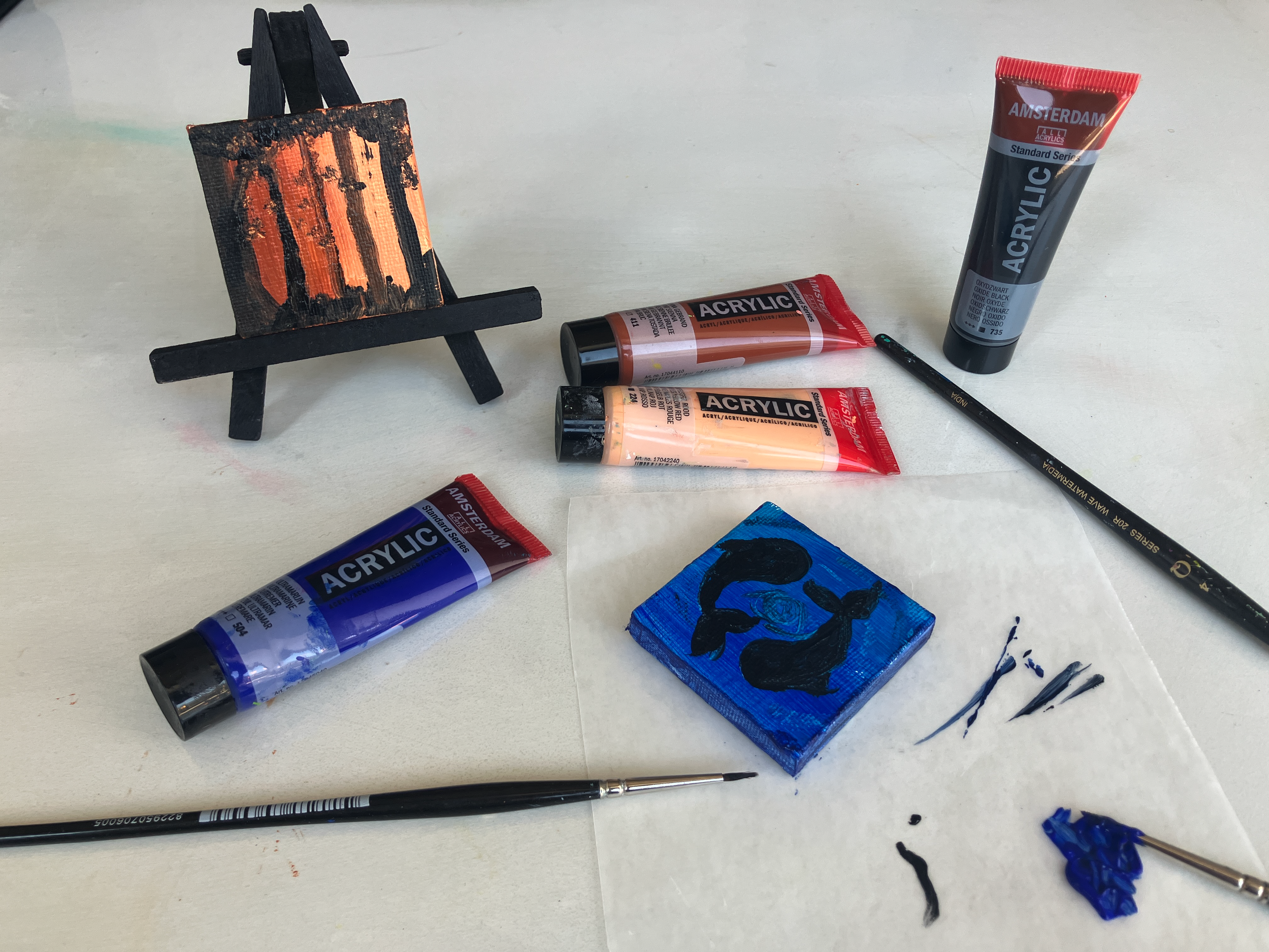 Small easel with a painted black square featuring orange stripes, surrounded by acrylic paint tubes in blue, peach, brown, and gray, two paintbrushes, and a blue block with dark shapes on a white surface.