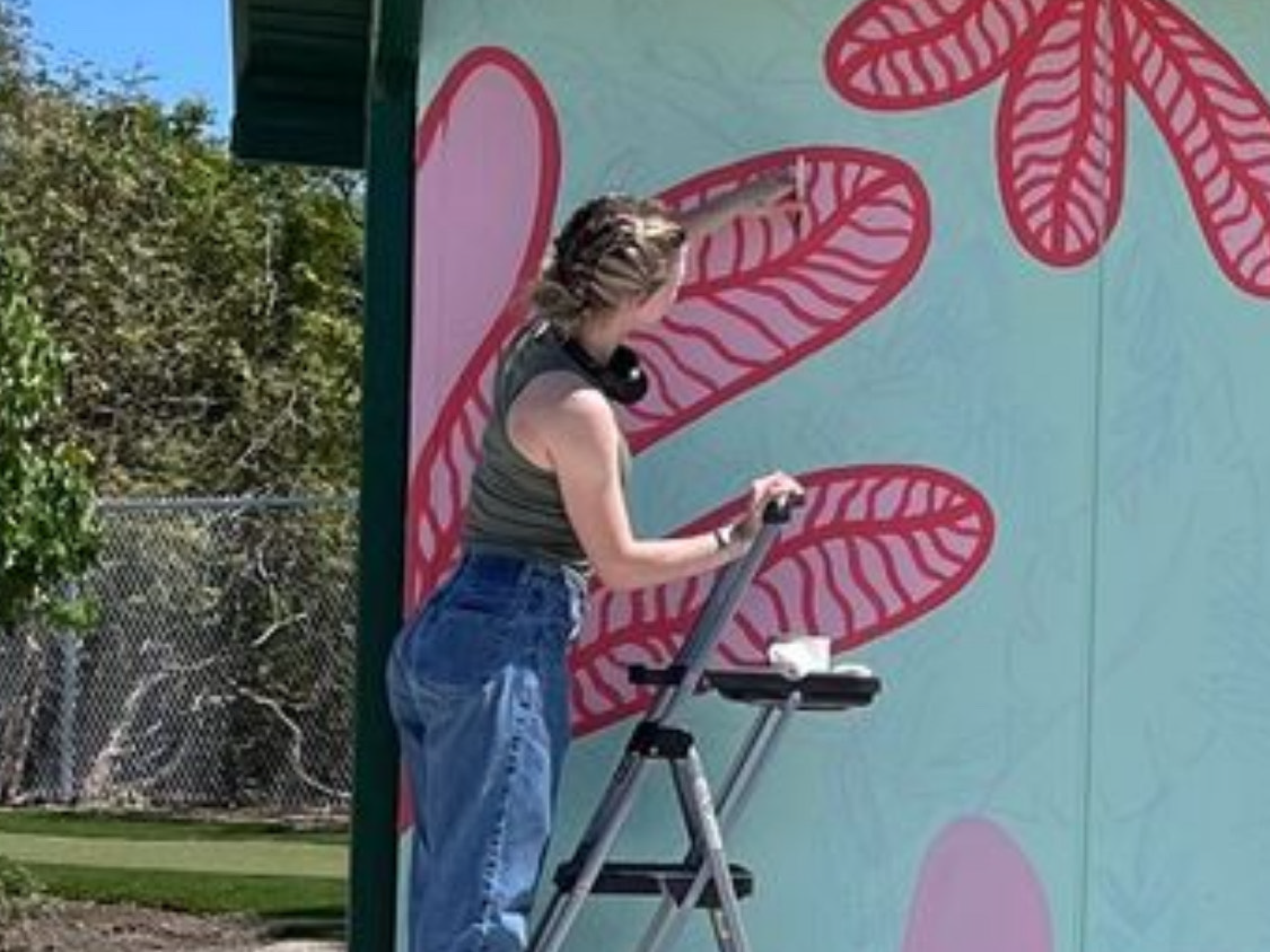 Person on a step ladder painting a large outdoor mural with red leaf‑like shapes on a pale teal wall.