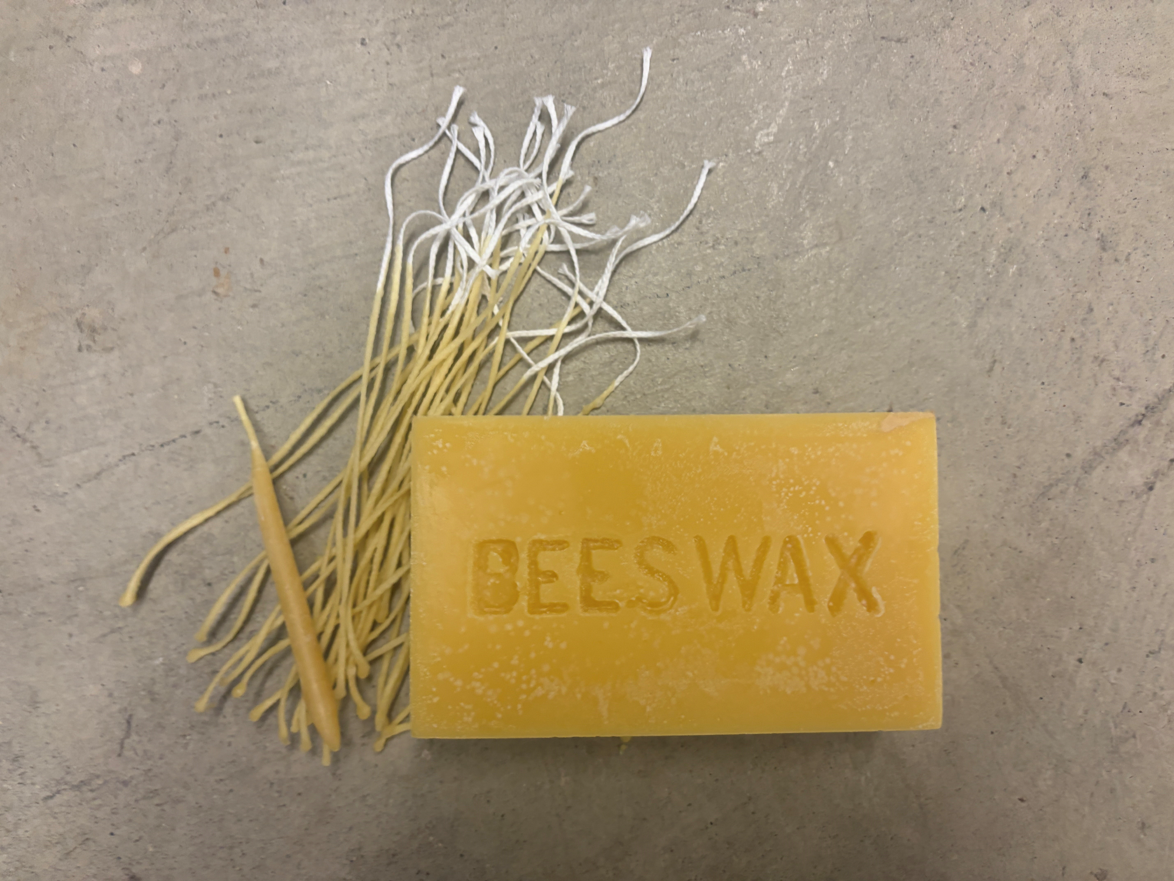 Beeswax block and bundle of candle wicks displayed on a concrete surface.