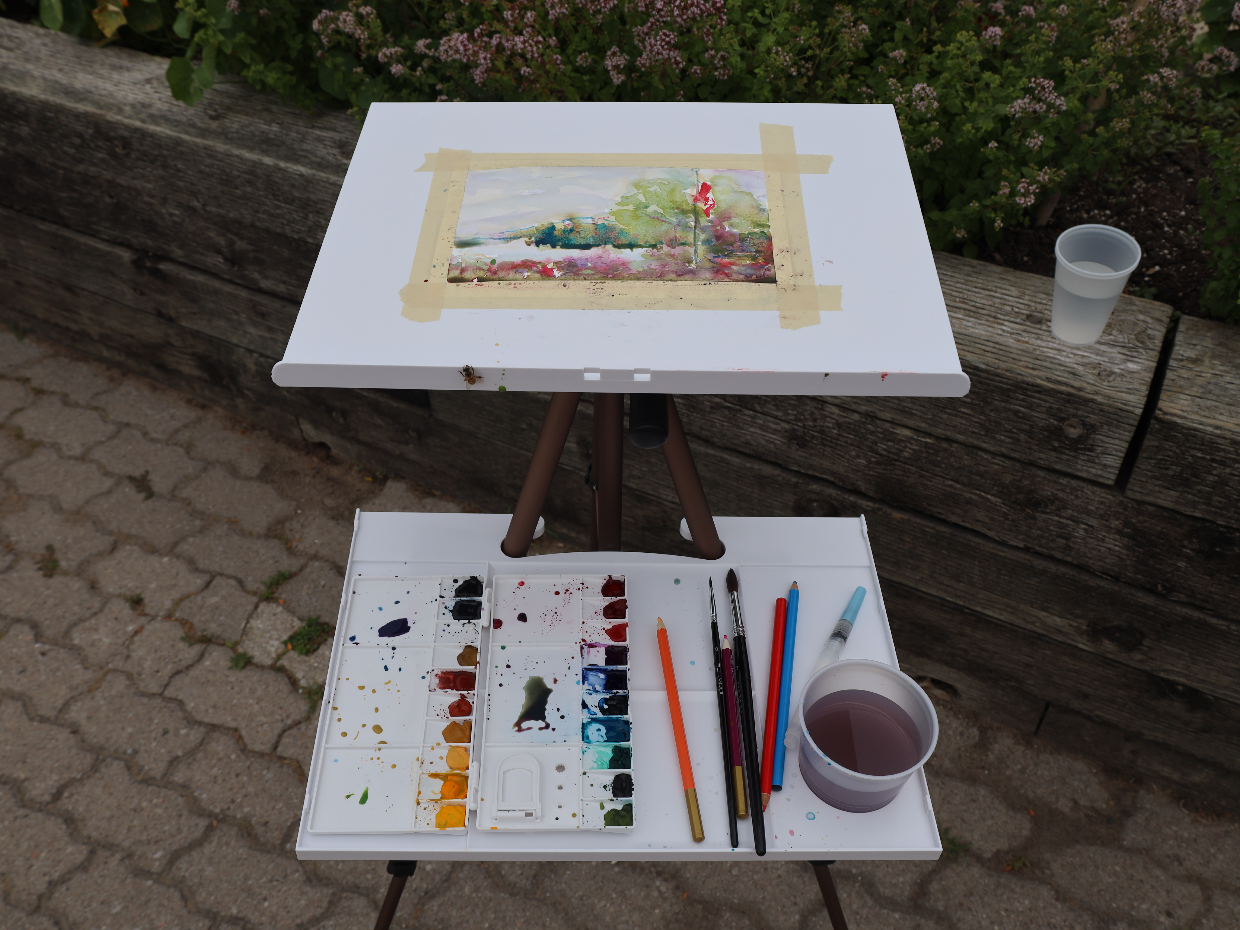 An outdoor painting setup with a watercolor palette, brushes, water cups, and a taped landscape painting on an easel.