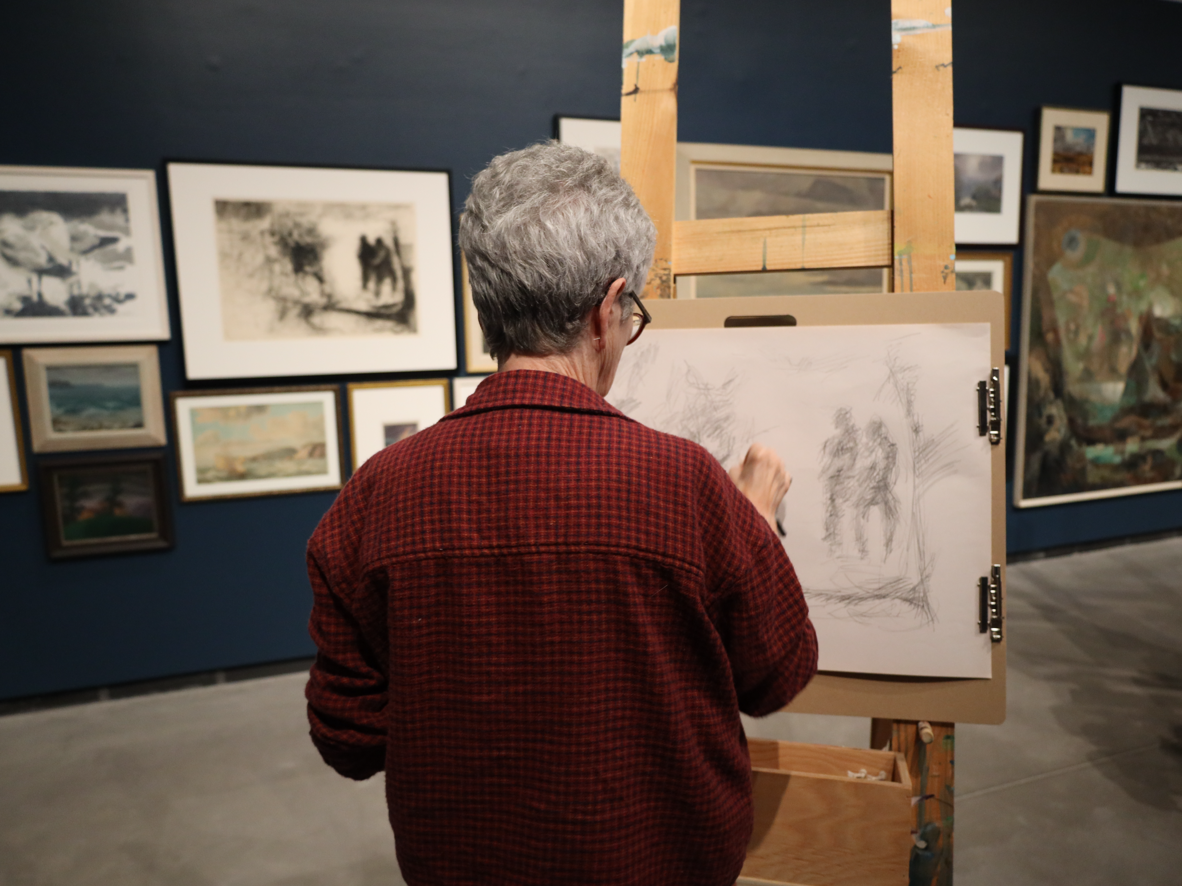 A person sketches on a large drawing board inside a gallery filled with framed artworks on the walls.