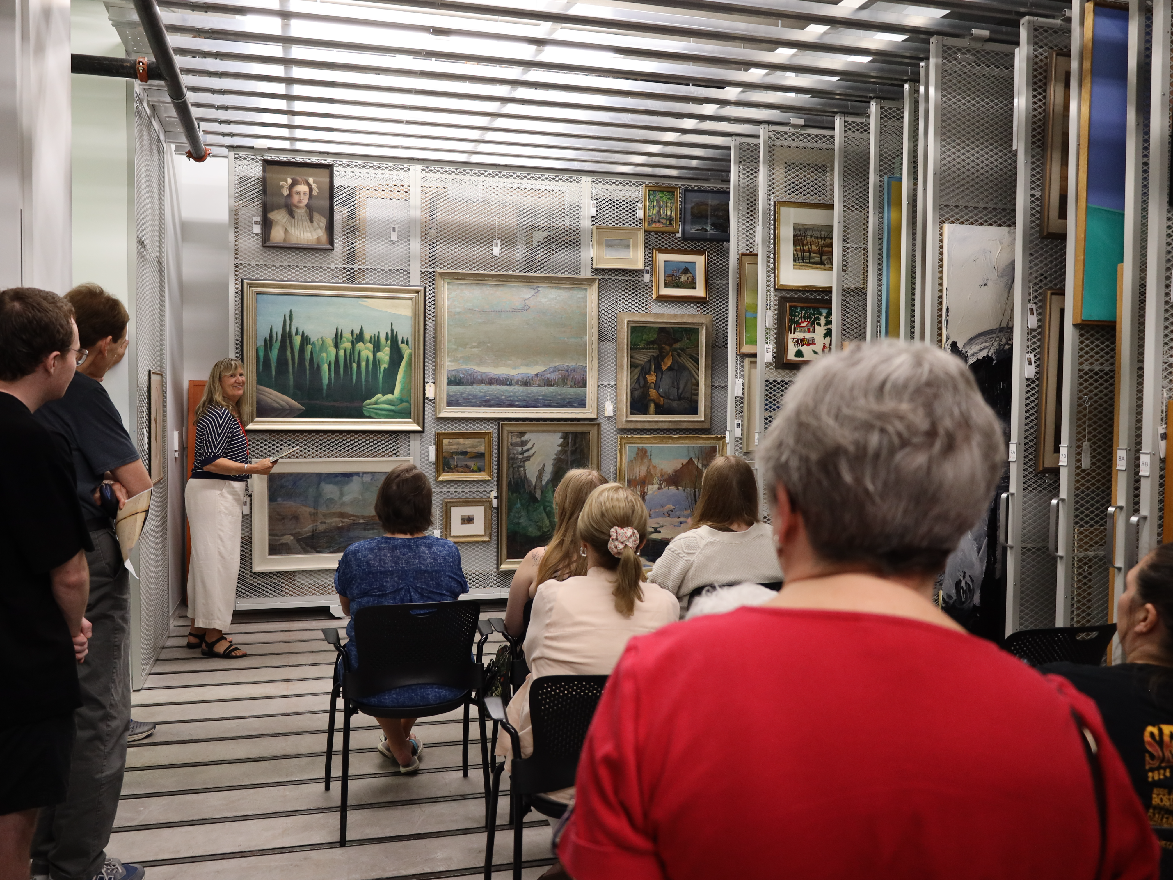 A group of seated visitors listens to a guide speaking in a storage area lined with framed paintings on sliding racks.