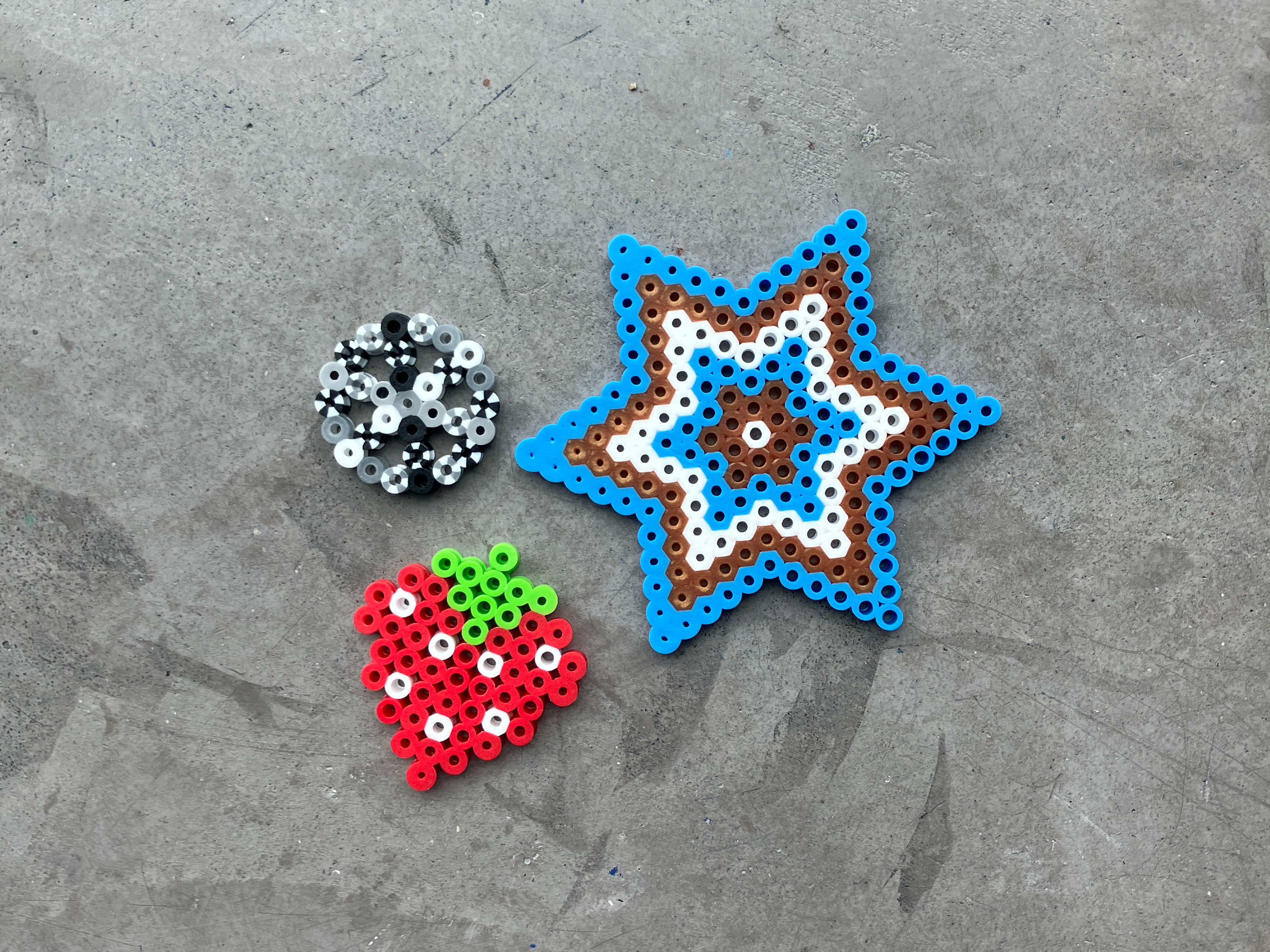 Perler beads in various colours, sit fuzed together to make the following shapes: A snowflake, a wheel, and a strawberry. They lay on top of a grey table.