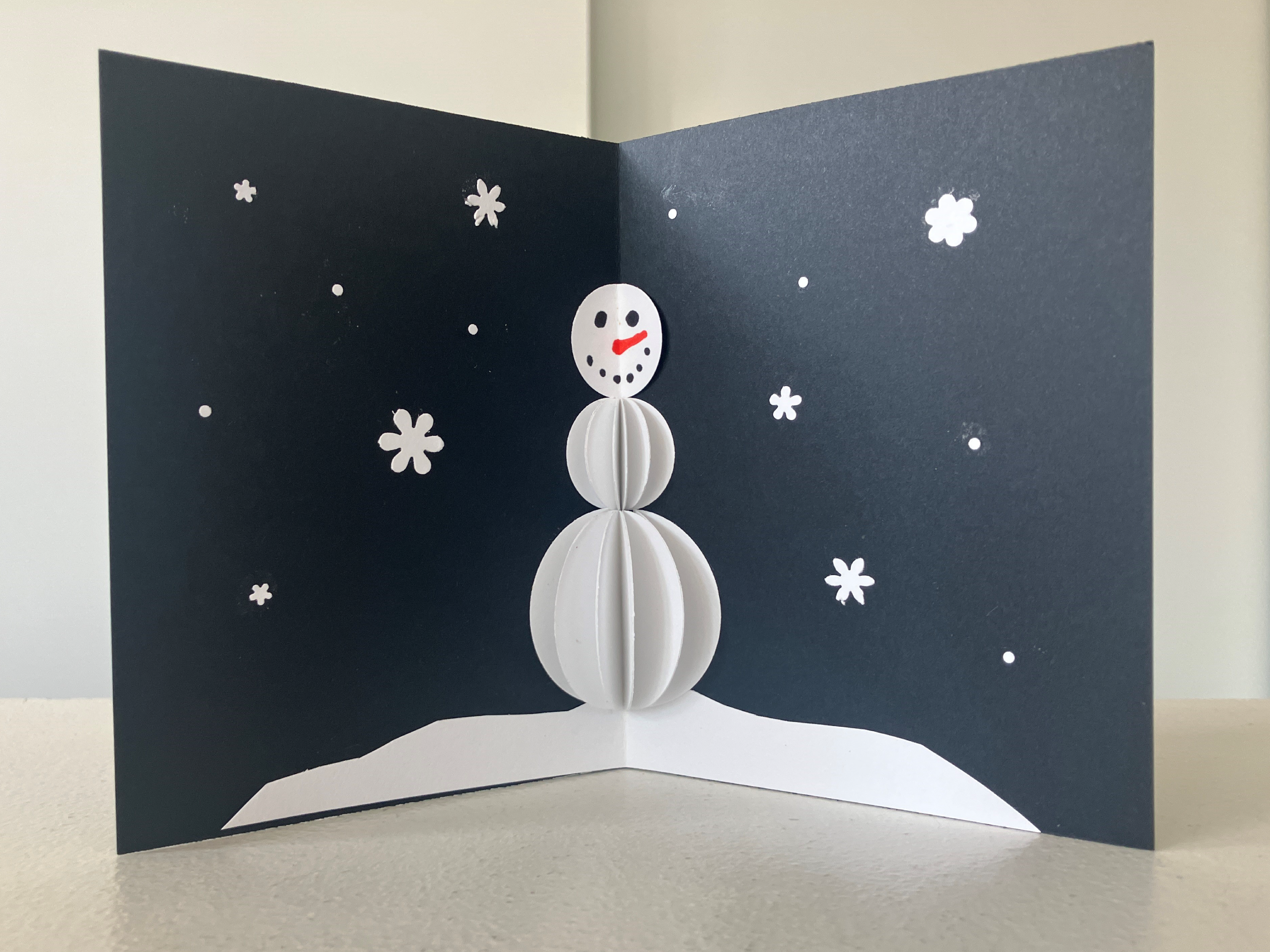 A 3-D style card sits on a grey table. The card is made from navy coloured paper and features a 3D snowman in the centerfold of the card. Little white snowflakes are also drawn around the snowman.