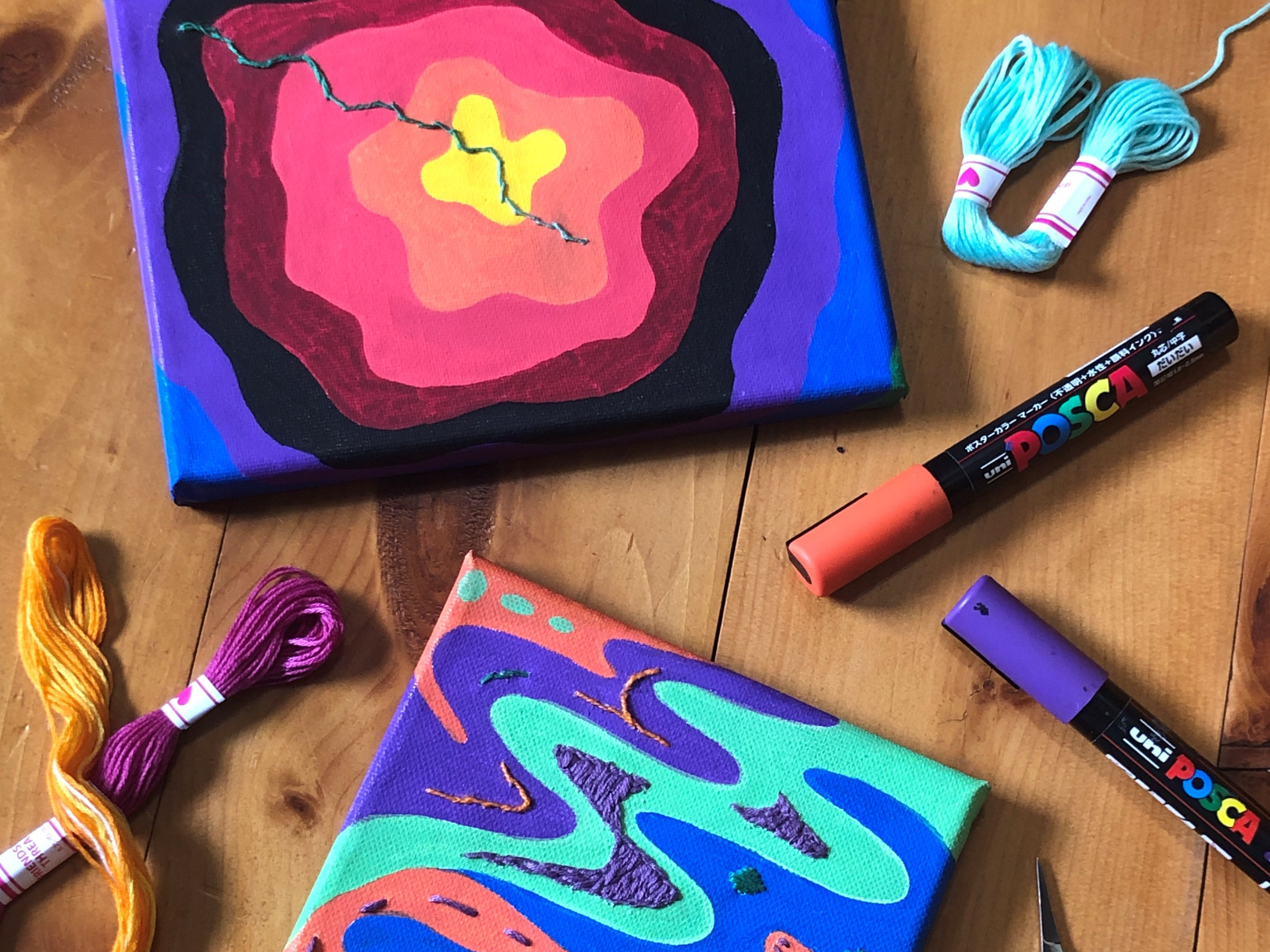 Vibrant painted and embroidered canvases surrounded by embroidery floss and paint markers.