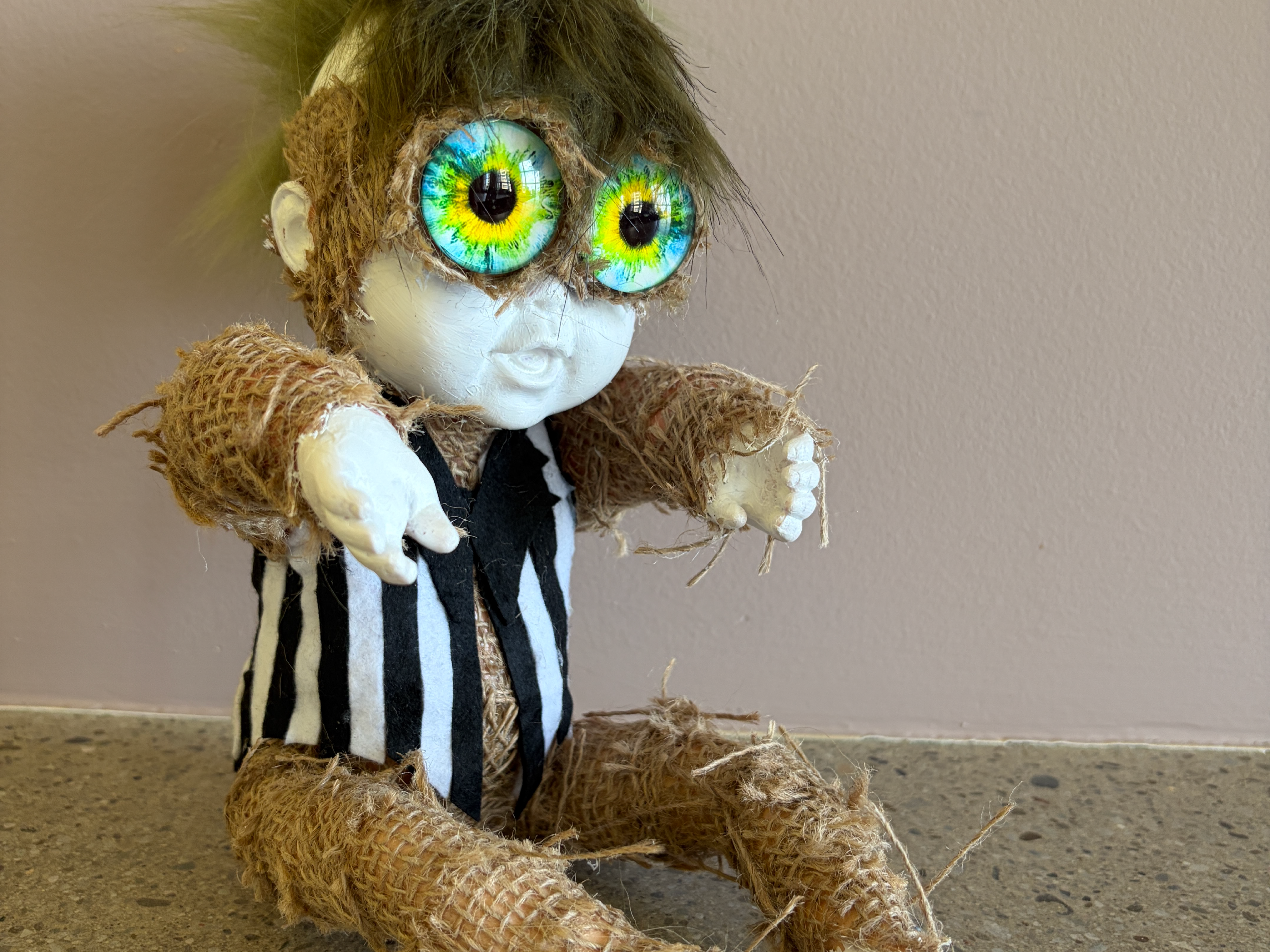 Handcrafted doll with oversized green eyes, striped scarf, and textured burlap clothing.