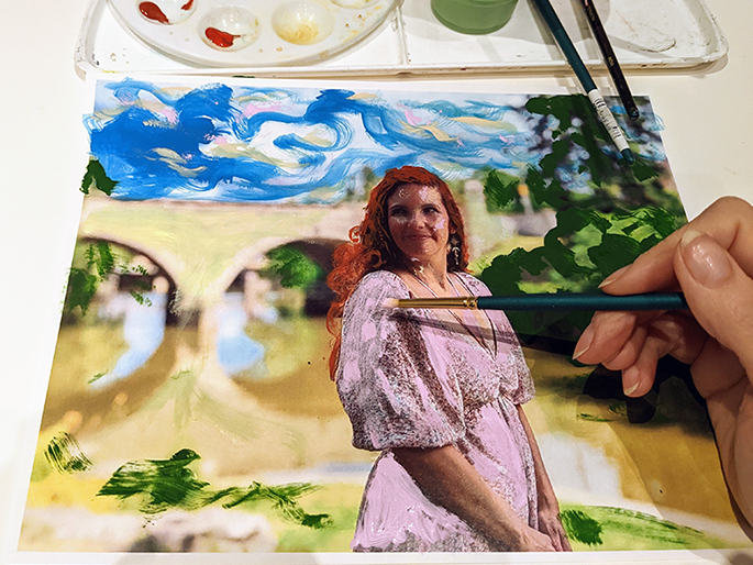 A photographic image of a woman with red hair and a pink dress standing in front of a acrylic painting of bridge.