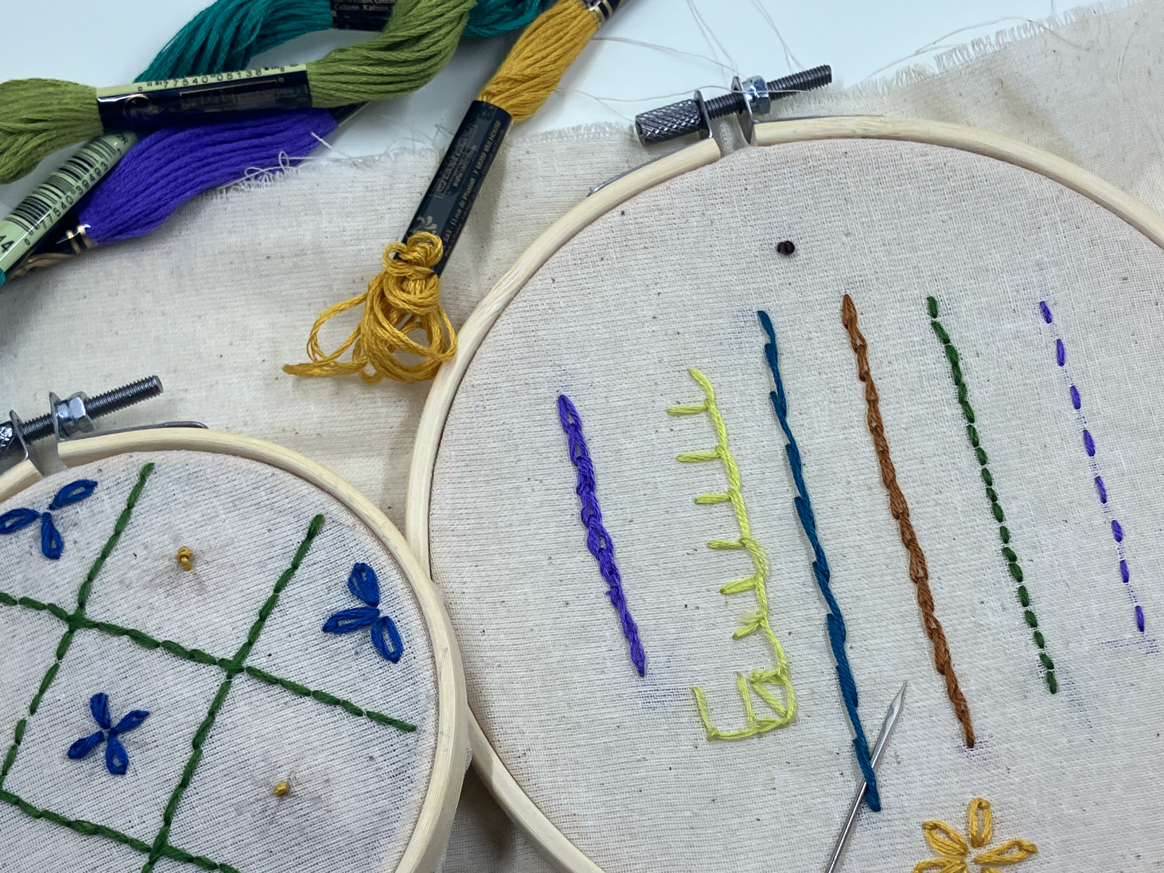 Two embroidery hoops, a needle, and different coloured embroidery thread sits on a table. In the embroidery hoops, different stitching techniques are visible on the fabric.