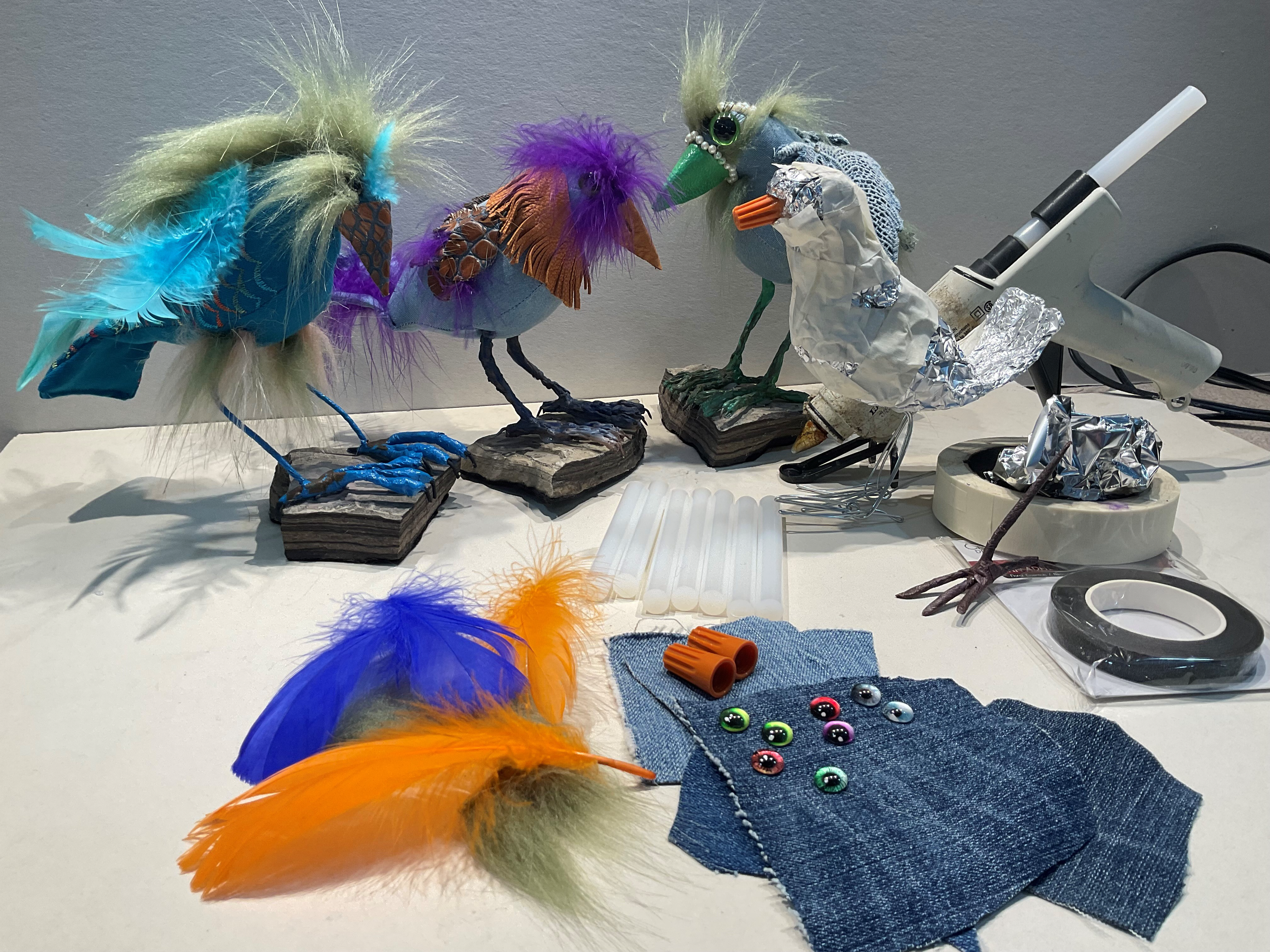 Three birds sculpted from tin foil and adorned with feathers and other material, sit on a table in front of various fabric scraps, a hot glue gun, and rolls of tape.