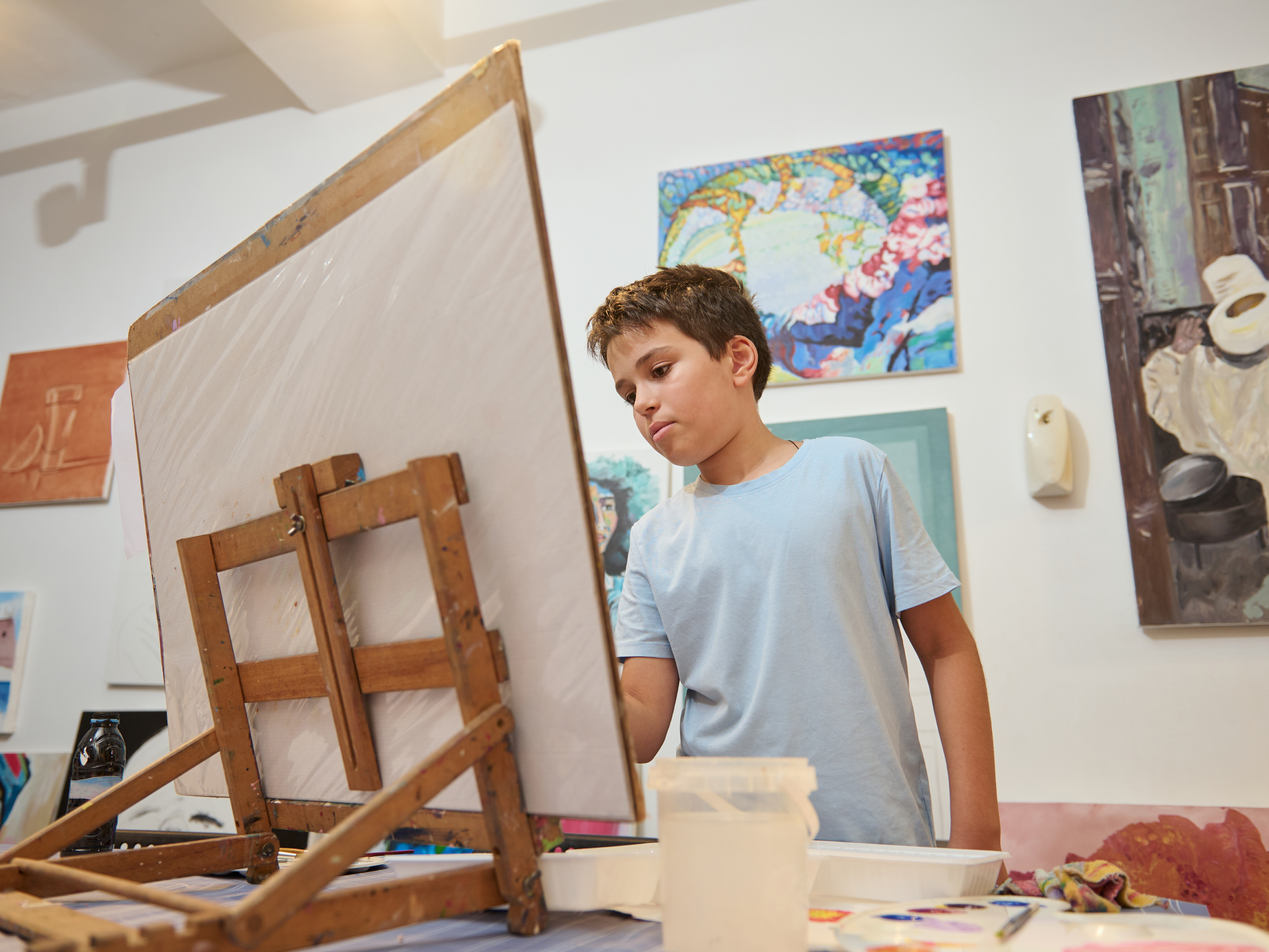 A young person painting at an easel in a studio filled with colourful artwork.