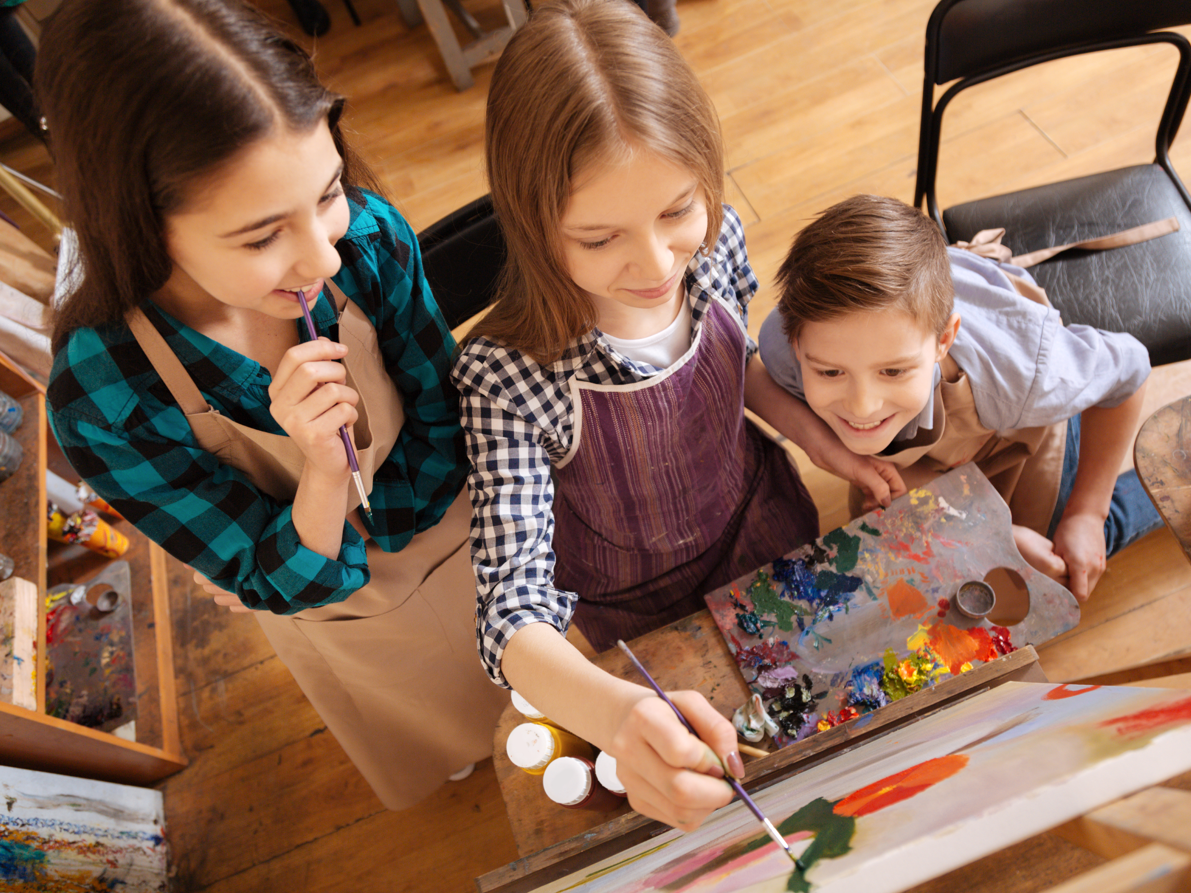 Three young participants paint at an easel, surrounded by brushes and a palette filled with bright colours.