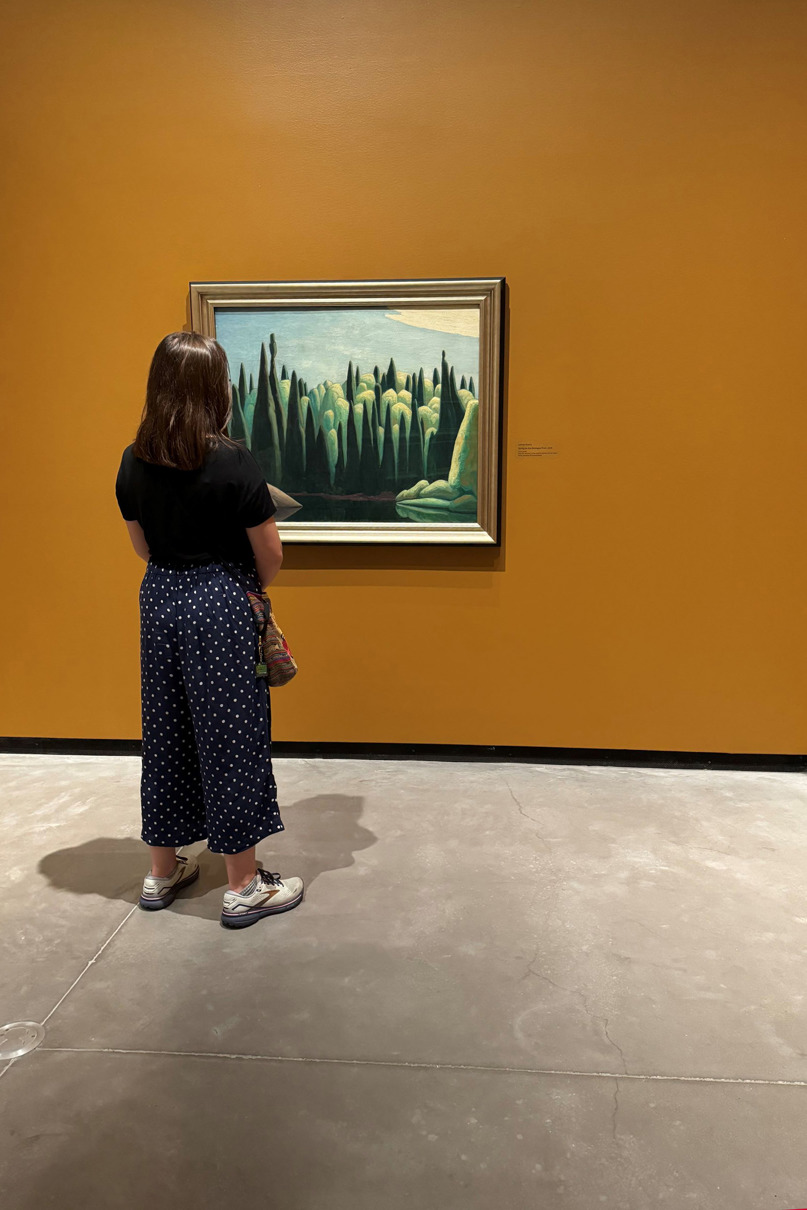 A woman with black shirt and blue polkadot pants looks at a painting of green trees and a blue sky hanging on an orange wall