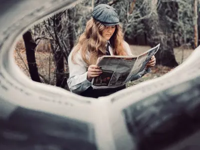 Humans of Lambton overall winner. A young girl wearing a flat cap is seen reading the paper. The photo is shot looking through a newspaper.