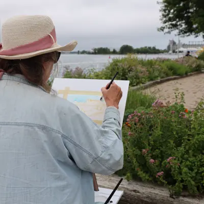 A woman dressed in a light blue shirt and a straw hat with pink ribbon, paints outside down at the Sarnia waterfront.